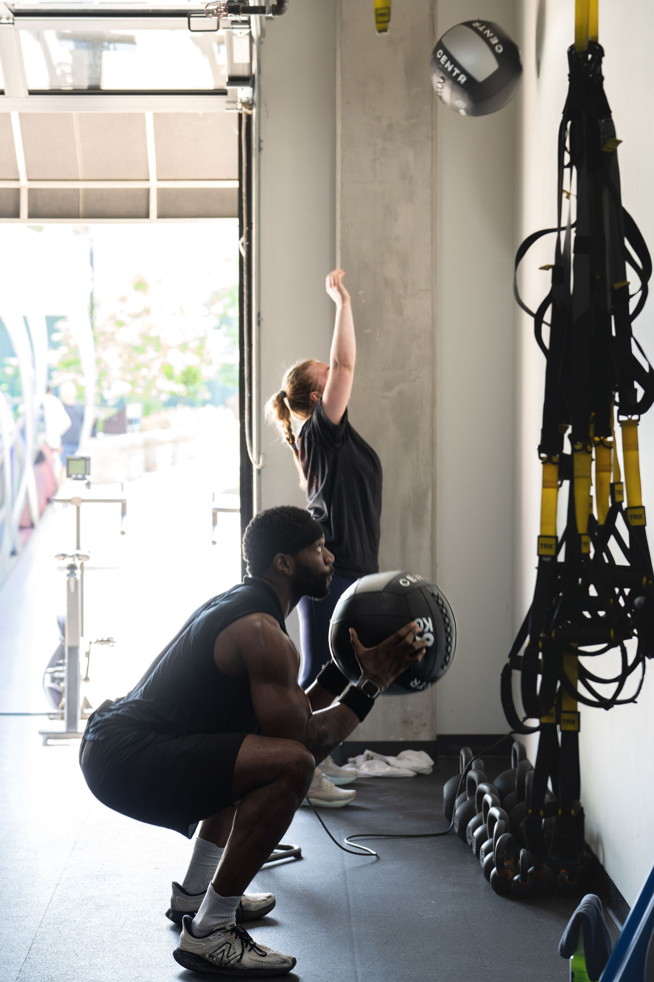 Two members doing wall-ball exercises in a PKFit gym beside an open bay door