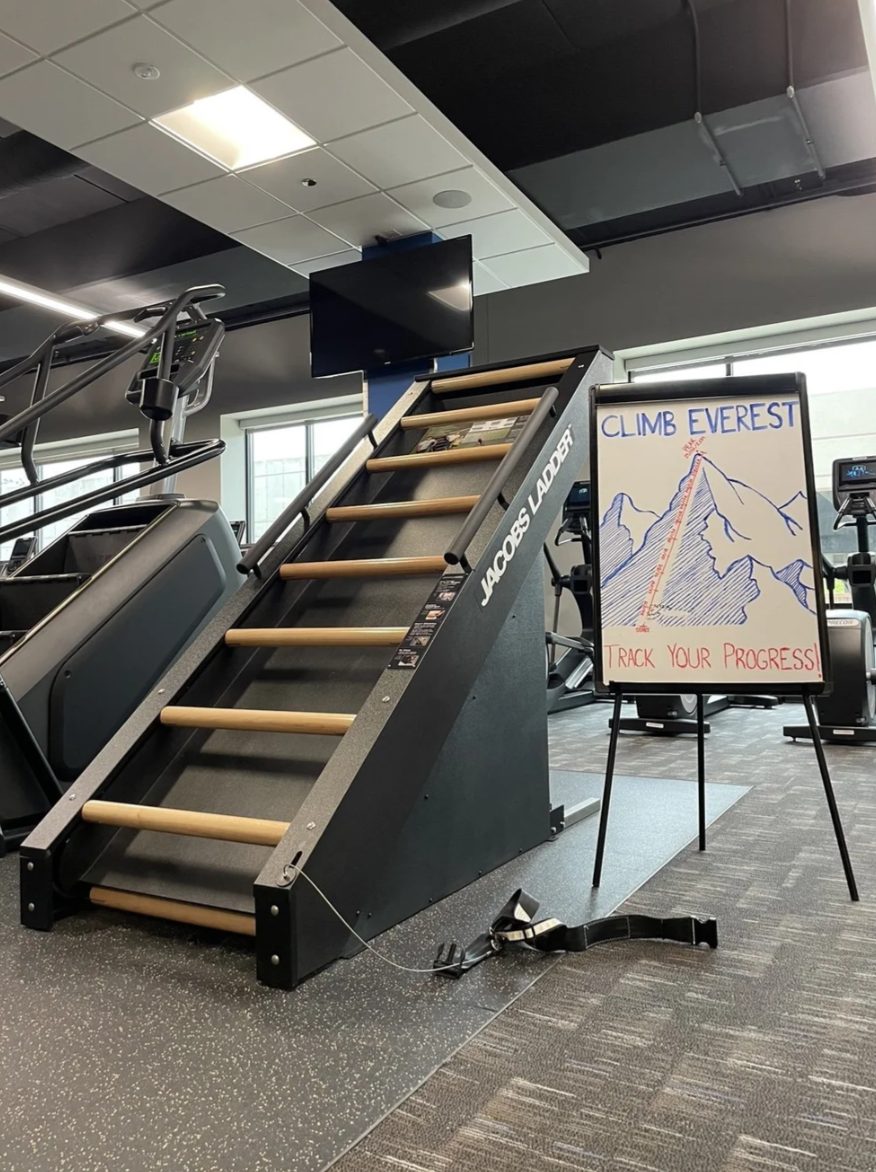 The Jacobs Ladder climbing machine at PKFit Madison East beside a Climb Everest progress whiteboard