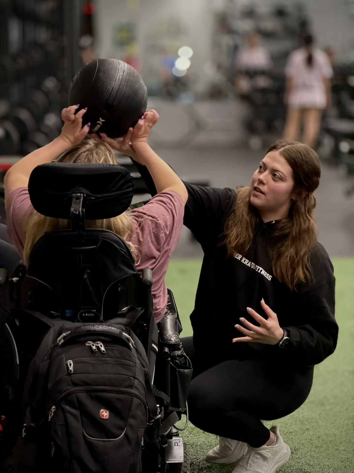 A PKFit trainer coaching a member in a wheelchair through a medicine ball lift
