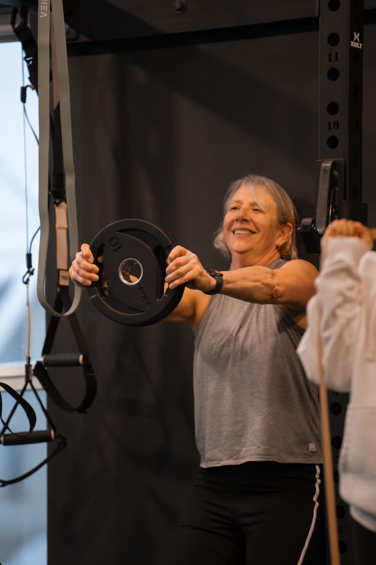 A smiling member holding a weight plate in front of her at a PKFit rack