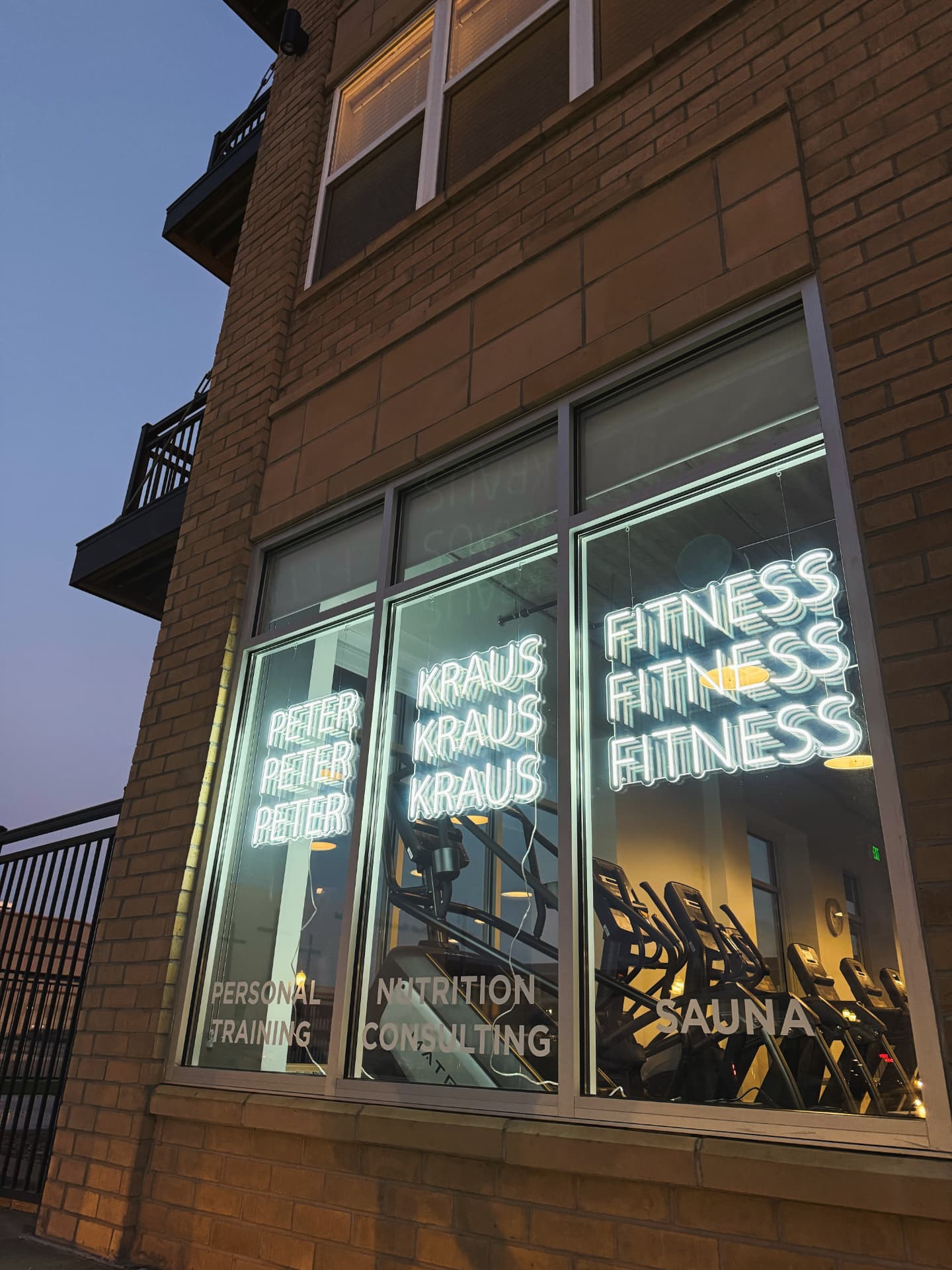 The PKFit Wausau storefront at dusk, with illuminated Peter Kraus Fitness window signs above the cardio machines