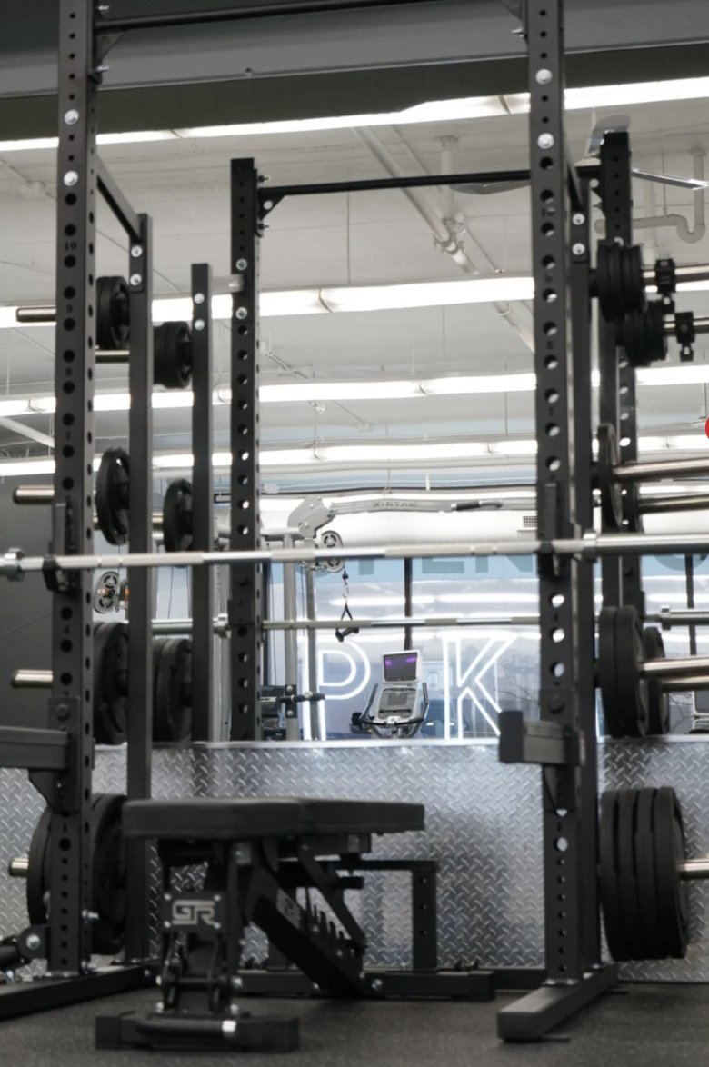 A squat rack at University Row with the PKFit neon sign reflected in the mirror