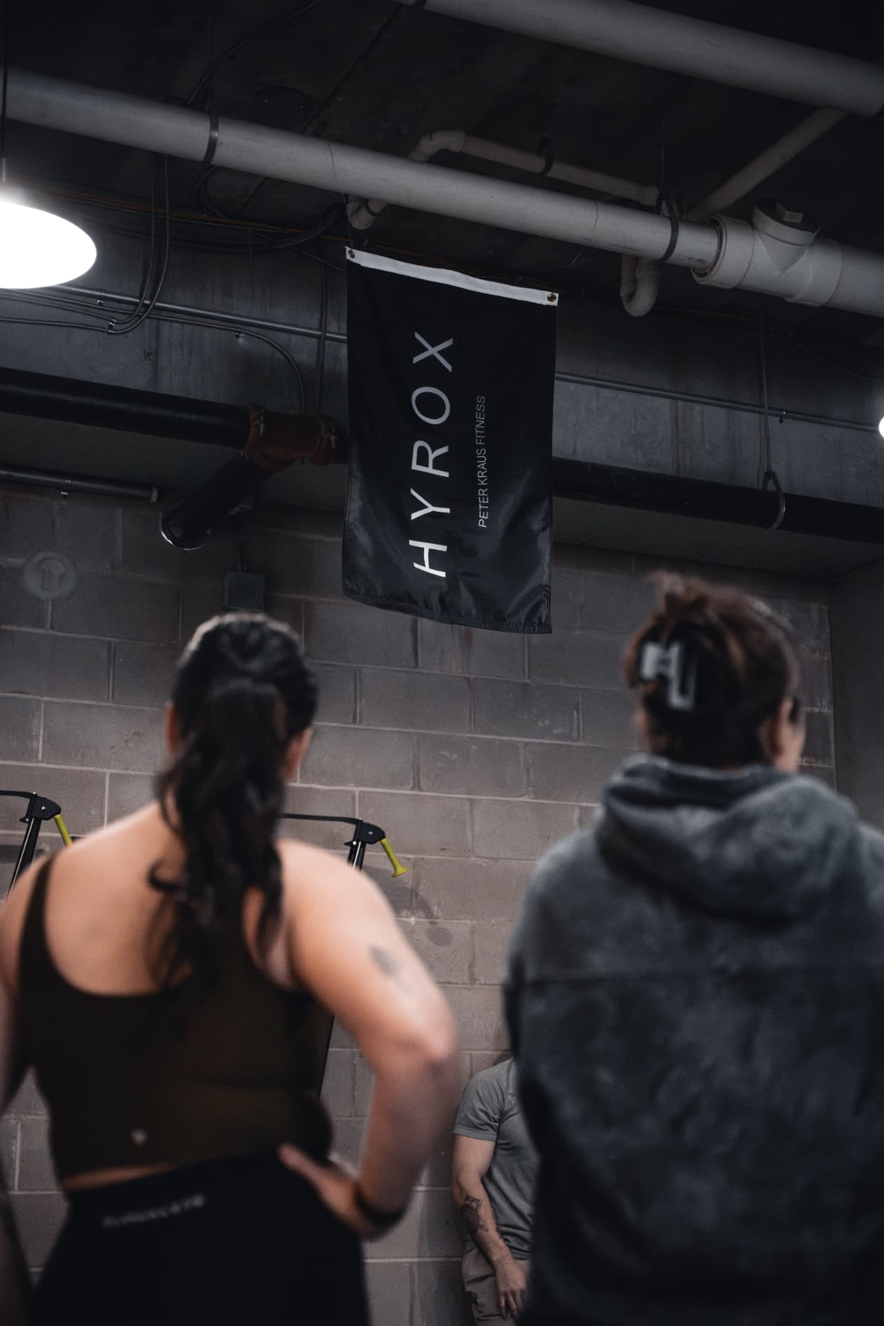 Two members facing the front of a class beneath a PKFit HYROX banner