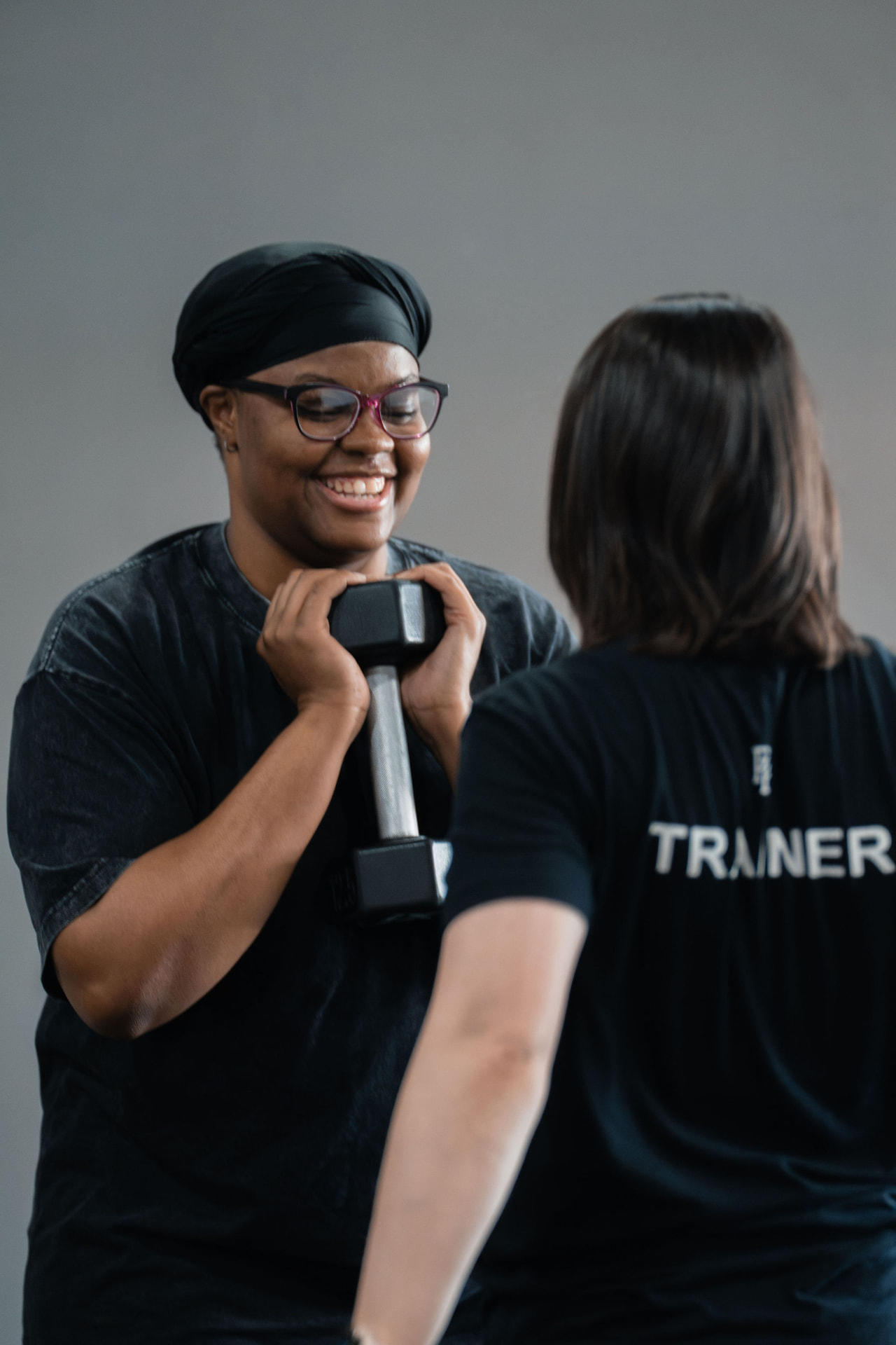 A smiling PKFit member holding a dumbbell while a trainer coaches her