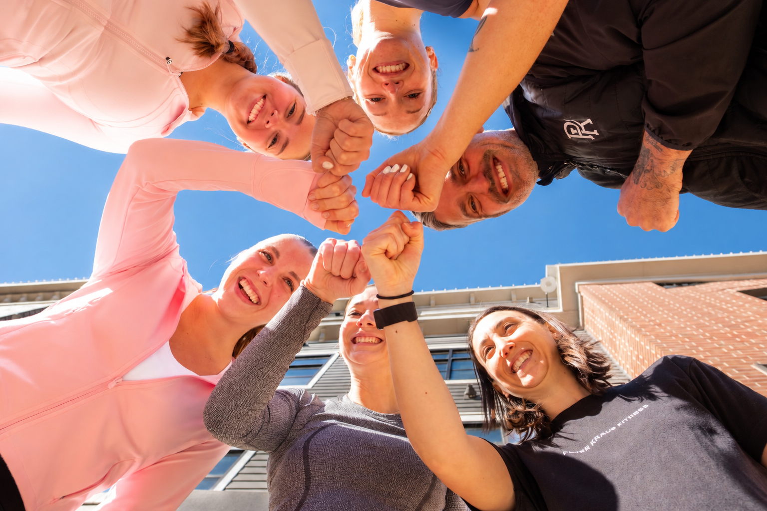 Six PKFit team members in a huddle with their hands stacked together, photographed from below against a blue sky